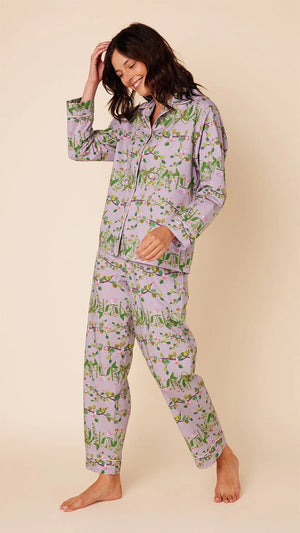 A woman stands against a plain background, smiling and touching her hair. She is wearing the CAT'S PAJAMAS - SAKURA Luxe Pima Cotton Pajamas in matching purple floral with green accents. The Pima Cotton pajamas from CAT'S PAJAMAS consist of a button-up long-sleeve top and pants. She is barefoot and appears relaxed and happy.