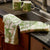 The CASKATA - Arbor Birds Kitchen Towel in Green features two folded fabric napkins with a green botanical print of birds on leafy branches over a white background, perfect as kitchen linens or charming Mother's Day gifts.