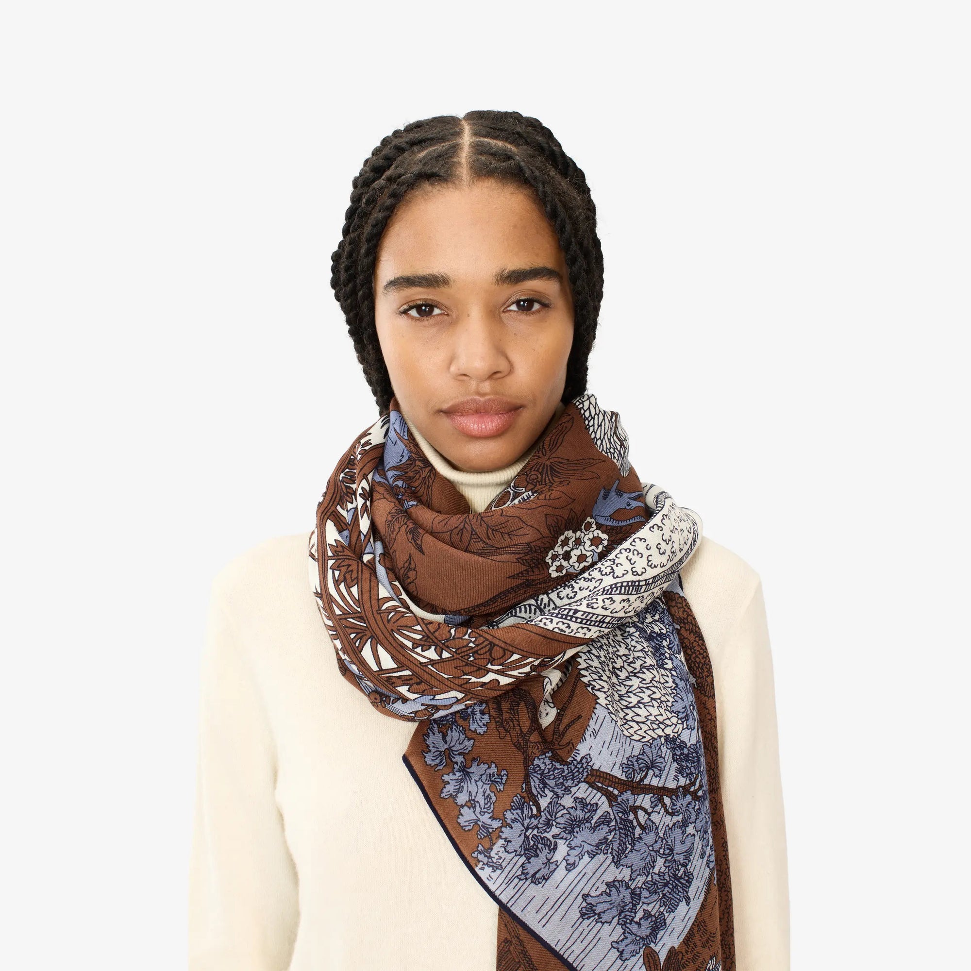 A person stands wearing the INOUI EDITIONS - QUATRE SAISONS SCARF, a long and lightweight summer accessory featuring detailed, intricate designs in brown, blue, and white tones, draped over a cream-colored top. The unique and colorful scarf from INOUI hangs elegantly around the neck and over one shoulder against a plain white background.