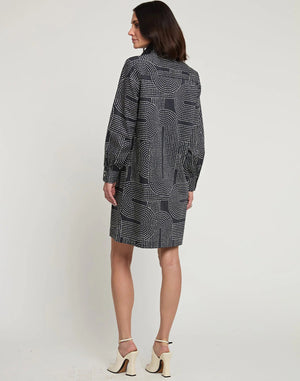 A woman with shoulder-length dark hair is wearing the new HINSON WU Vicky Long Sleeve Dress with Saddle Stitch Print, a knee-length outfit featuring a geometric dot pattern in shades of grey and white. Paired with white high-heeled sandals, she stands against a plain, off-white background.