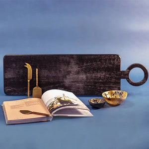 A black cutting board with a round handle displays three gold cheese knives of different shapes. Next to it are two Medium Horn Bowls by Be Home, Inc., speckled in brown, and a hardcover book titled "Platters and Boards" showcasing a colorful food spread on the cover—a perfect serving kit that's ethically crafted.