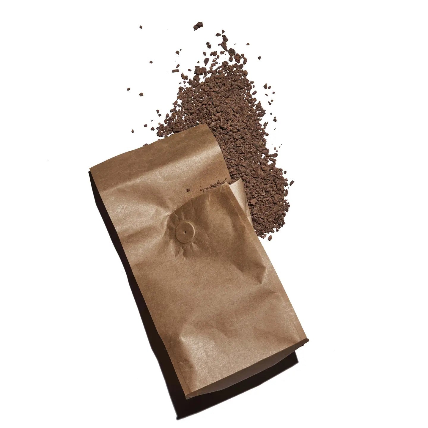 A crumpled brown paper bag lies on a white surface with coffee grounds spilling out of it. The espresso fragments gather in a small pile beside the bag, evocative of how RITUAL CHOCOLATE - ONE HUNDRED PERCENT DRINKING CHOCOLATE shavings might scatter when unwrapped.