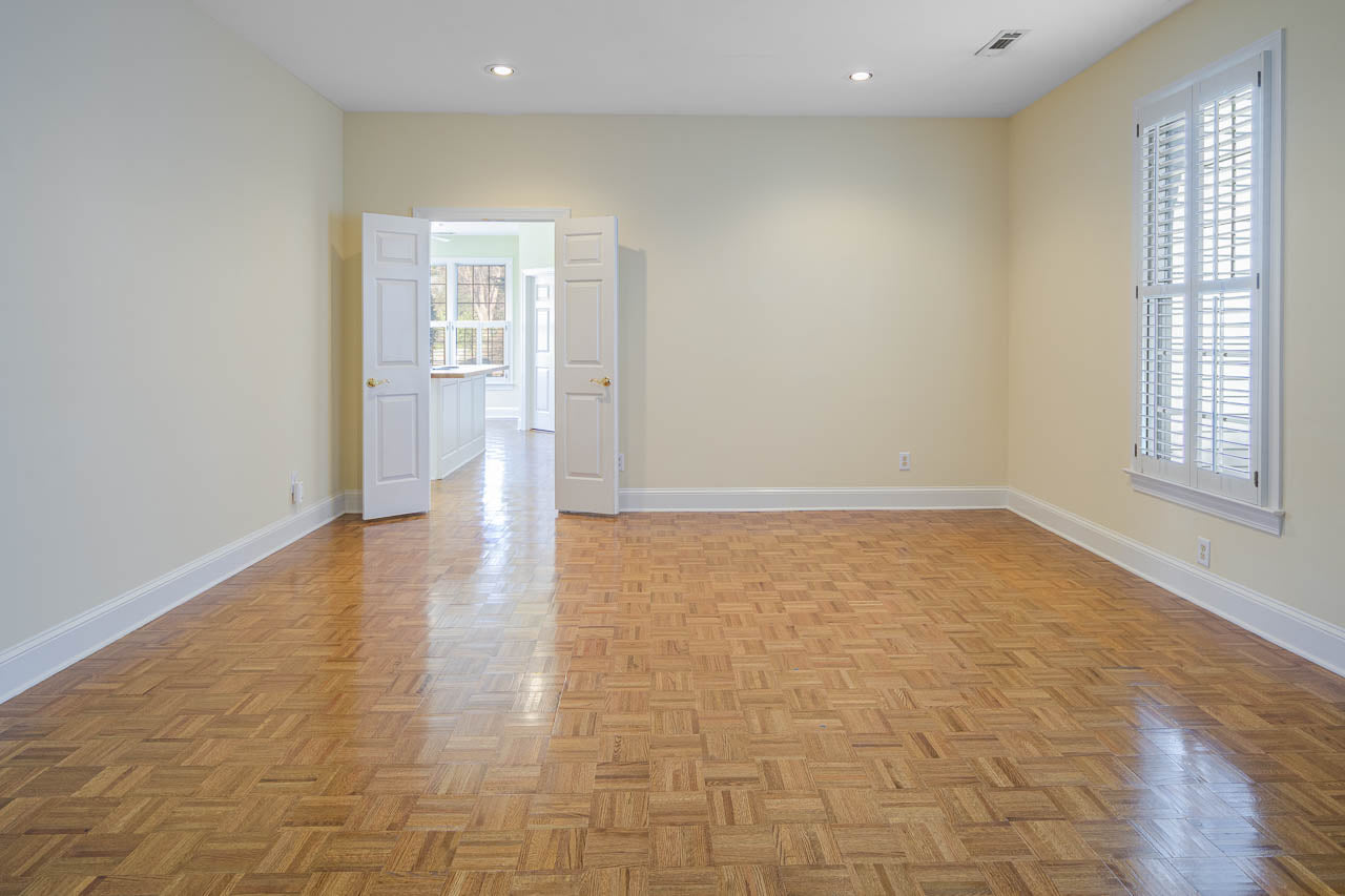 Empty room with light beige walls, parquet wood flooring, white trim, and white double doors slightly open. Sunlight streams through a window with white shutters on the right wall. Ceiling lights are recessed.