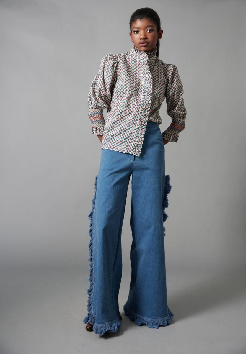 A woman models the HOPE FOR FLOWERS - FRILLED BUTTON DOWN SHIRT by HOPE FOR FLOWERS, made from GOTS certified organic cotton, with a high collar and wide sleeves, paired with flared blue jeans featuring fringe along the side seams.