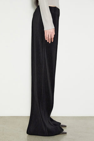 Side view of a person wearing the MEIMEIJ Long Pleated Pant in black, highlighting the elegant texture of pleated fabric. The pants are paired with a light gray, long-sleeved top, and the ensemble's draping is well highlighted against a plain background and light-colored floor.