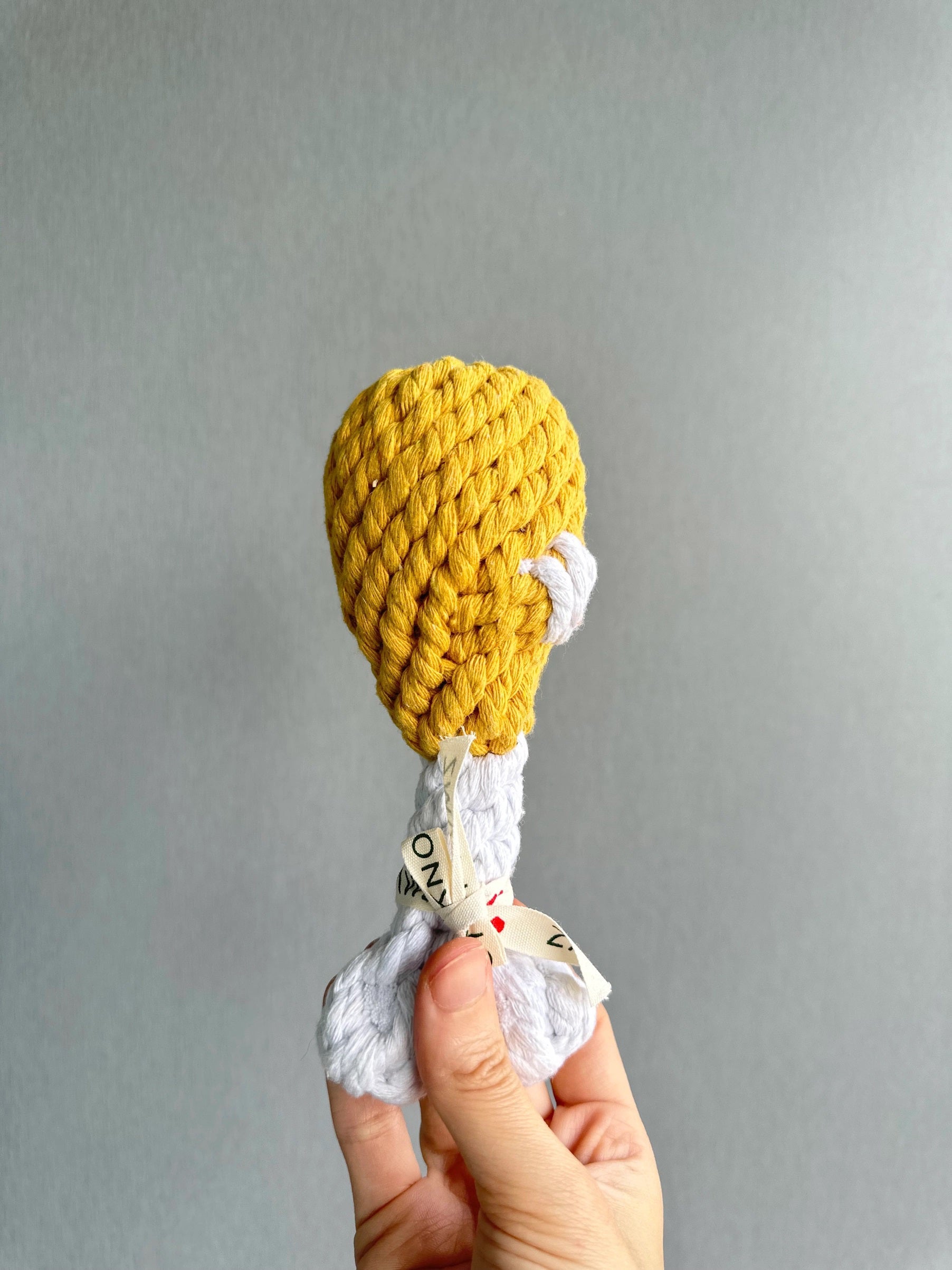 A hand holds the KNOTTY PAWS - CLUCKY DRUMSTICK DOG TOY by KNOTTY PAWZ, resembling a drumstick with yellow yarn as the fried top and white/brown fabric as the bone. Perfect for pups!.