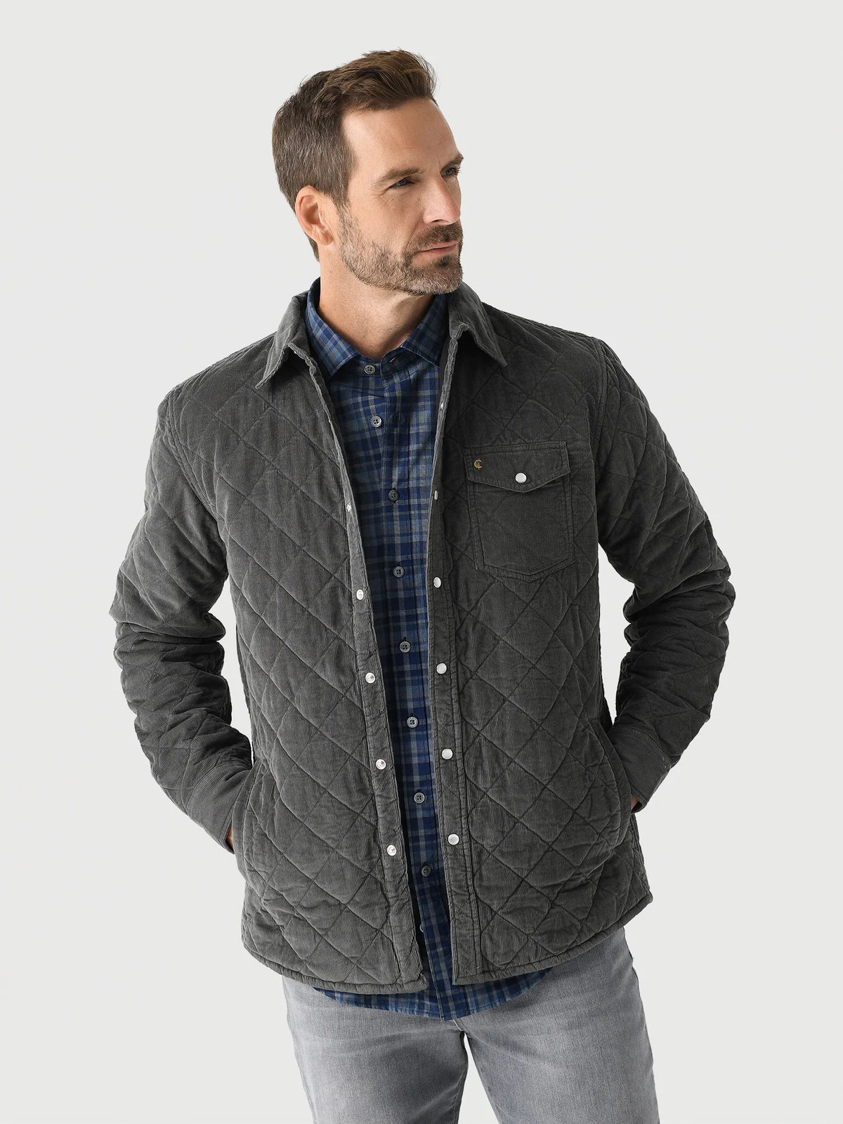 A man with short, curly hair and a beard wears the CRIQUET - CORDUROY QUILTED SHACKET by CRIQUET and blue jeans, standing against a white background while smiling and looking to his right.