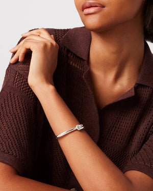 A woman in a brown knit sweater wears a gold Maeve Bangle by JENNYBIRD, featuring circular details, as she looks sideways. Her left hand rests on her opposite shoulder against a plain light gray background.