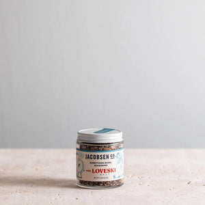 A small glass jar of Jacobsen Salt Co. Everything Bagel Seasoning, a collaboration with Loveski, sits on a flat surface against a plain light gray background. The jar's label is white with blue and red text and features cartoon illustrations. Perfect for your homemade sourdough bagels, this blend of seasonings stems from Northern California's artisanal traditions.