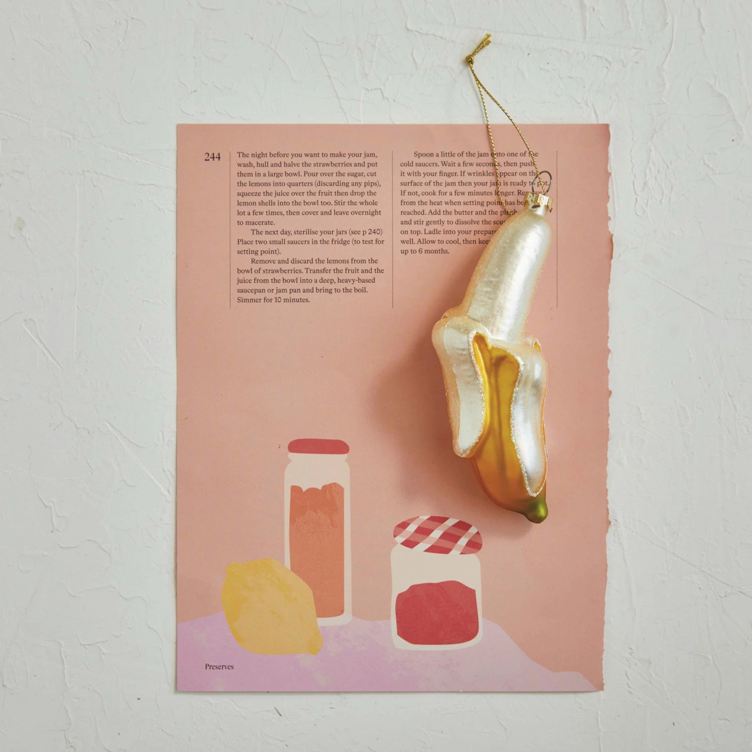 A page showcasing illustrated jars and a lemon is set against a peach background in front of a textured white wall. Hanging in the top right corner, the GLASS PEELED BANANA ORNAMENT from CREATIVE COOP features a glittering finish with a gold string, enhancing the elegance of the yellow & cream color scheme.