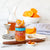 A jar of Stonewall Kitchen - Tillen Farms Candied Orange Peels is placed on a marble counter next to a cocktail glass, sliced oranges, and a filled carafe. A jigger, cocktail strainer, and spoon complete this vegan-friendly citrusy treat preparation scene.