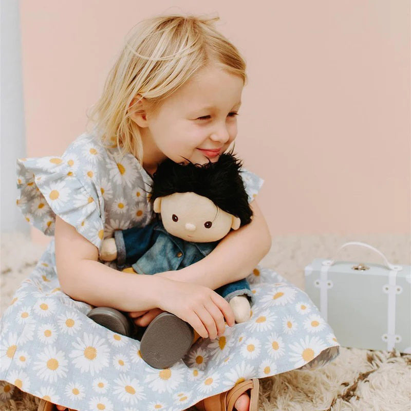 A Dinkum Doll from Olli Ella USA&#39;s Travel Togs collection, featuring dark hair and dressed in a denim jacket, mustard pants, and black shoes, stands on a wooden surface. Beside the doll is a teal See-Ya Suitcase with white trim and hardware. In the background, another larger teal suitcase is partially visible.