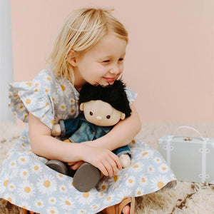 A Dinkum Doll from Olli Ella USA's Travel Togs collection, featuring dark hair and dressed in a denim jacket, mustard pants, and black shoes, stands on a wooden surface. Beside the doll is a teal See-Ya Suitcase with white trim and hardware. In the background, another larger teal suitcase is partially visible.