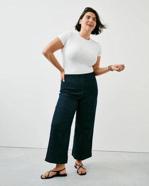 A woman is seen from behind, wearing a white sleeveless crop top and dark high-waisted SPANX - EVERYWEAR CROPPED WIDE LEG DENIM. Her hands are placed on her hips, with her blonde hair falling over one shoulder. The plain light gray background perfectly highlights her stylish SPANX denim that offers impeccable shape.