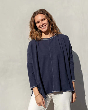 A person stands against a plain white wall, looking at the camera. They have curly hair pulled back and are wearing a loose-fitting, dark gray MERSEA Catalina Slub Long Sleeve Tee by MER SEA with 3/4 length sleeves, blue jeans, and a gold necklace with a circular pendant. Their shadow is visible on the wall behind them.