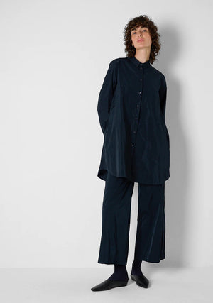 A person with curly hair wears the KATHARINA HOVMAN oversized taffeta blouse, featuring mother-of-pearl buttons, along with black pants. They stand in a minimalist setting with hands in pockets, gazing at the camera.