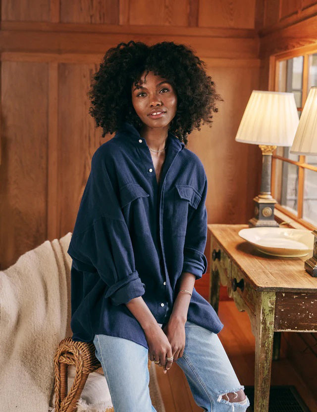 A navy blue, oversized utility jacket with long sleeves, folded at the cuffs. The FRANK AND EILEEN - MCLOGHLIN UTILITY JACKET features chest flap pockets and a label with text inside the collar. It is displayed on a white textured background, exuding that effortless California-cool layer vibe.