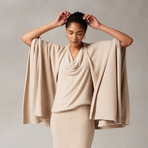 A person wearing the KASHWERE - TRAVEL PONCHO, a beige, draped poncho with a cowl neck and wide sleeves, poses with their arms raised and hands touching the back of their head. They have a neutral expression and are standing against a plain background.