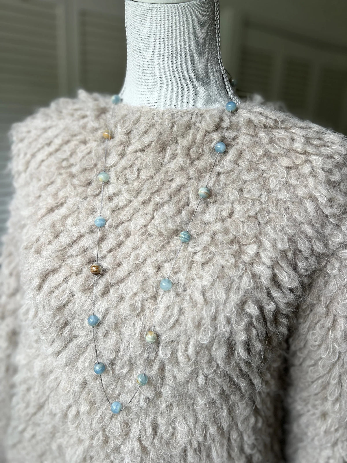 A mannequin in a beige textured sweater showcases the ANN LIGHTFOOT - BLUE CALCITE INTERMITTENT STRAND NECKLACE, a delicate 34&quot; piece with spaced blue and brown beads on a clear strand. The softly blurred background highlights the jewelry.