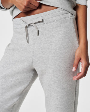 A person stands against a white background, smiling and looking down with one hand touching their hair. They are wearing a matching light beige SPANX AIRESSENTIALS TAPERED PANT and sweatshirt set made from ultra drapey, lightweight spacer fabric, paired with white sneakers.