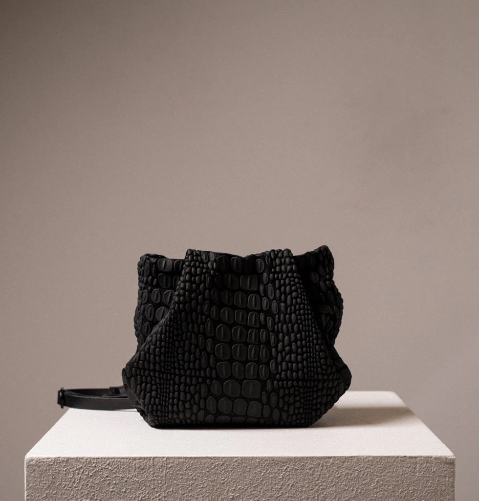 A black, textured handbag with a rectangular base and a gathered top is displayed on a white pedestal. The handbag, designed by DANIELA LEHAVI and named the TOKYO BALI BAG, features an alligator-like pattern and appears to be made of leather or a similar material. The background is plain and neutral-colored.