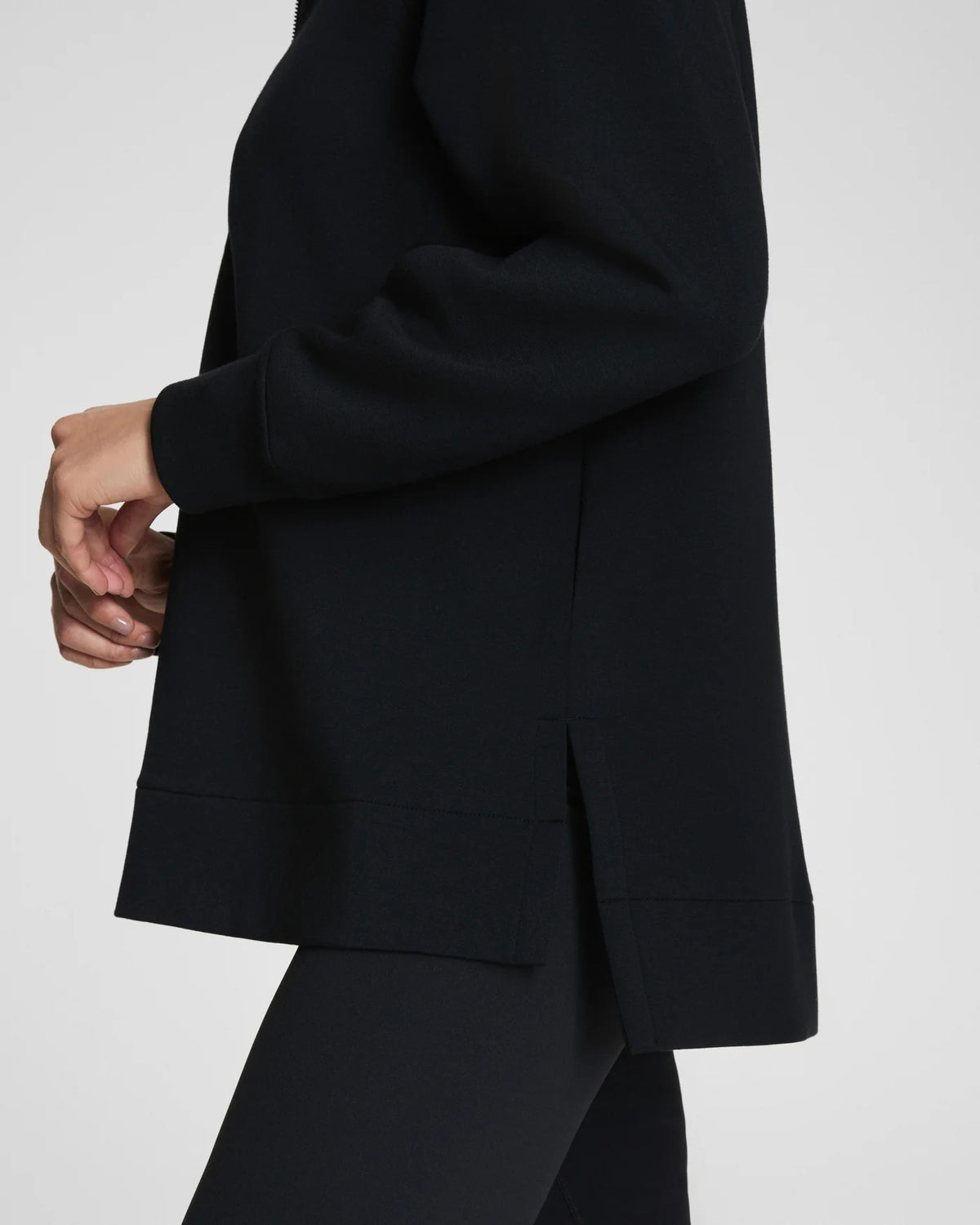 A woman with long dark hair stands confidently with arms crossed, wearing the SPANX - AirEssentials Brushed Half Zip Tunic Top in Black and matching leggings against a plain light gray background, showcasing stylish loungewear.