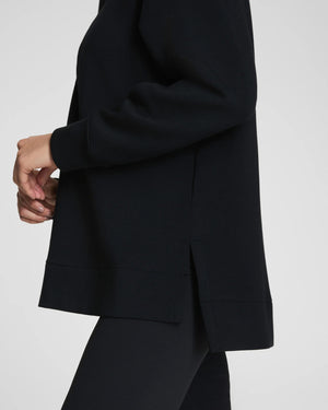 A woman with long dark hair stands confidently with arms crossed, wearing the SPANX - AirEssentials Brushed Half Zip Tunic Top in Black and matching leggings against a plain light gray background, showcasing stylish loungewear.