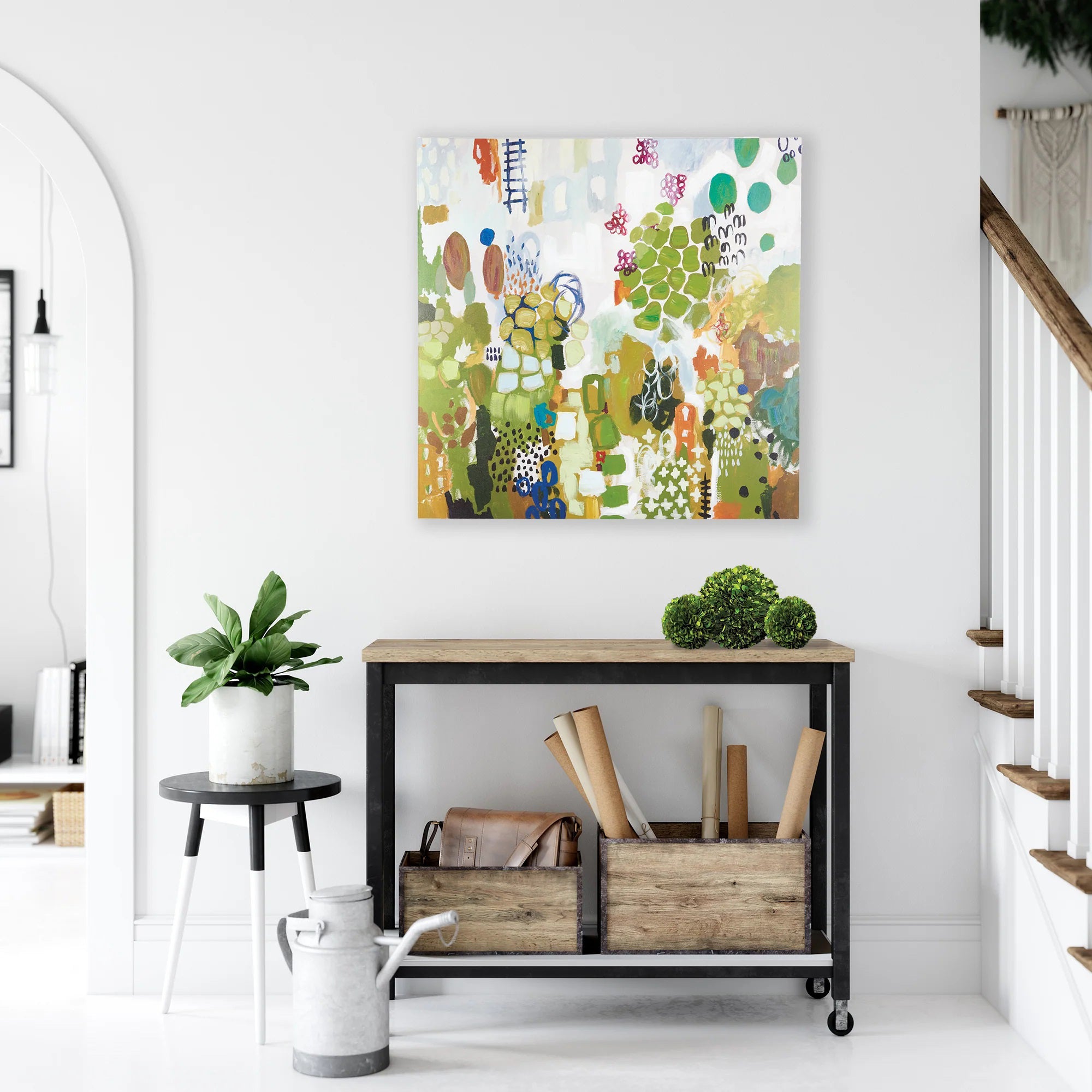 A bright, modern room displays the NAPA HOME AND GARDEN - SHANGRI LA ART on a white wall. Below it, a console table holds a plant, decor items, and rolled-up papers. A staircase with wooden steps is visible on the right, and an arched doorway completes the scene on the left.