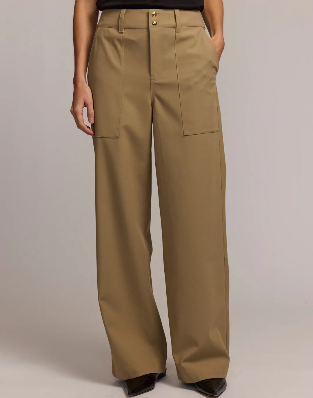 A person stands facing away, wearing HINSON WU&#39;s KELLY TRAVELER&#39;S TWILL PANT—high-waisted, wide-leg tan trousers with large back pockets—paired with a black top against a light neutral background.