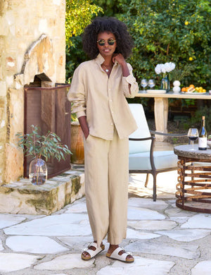 A woman with curly hair, wearing sunglasses, a beige long-sleeve shirt, and a matching pair of FRANK & EILEEN Maisie Wide Leg Linen Pull On Pants from the Tuscany Getaway Set, stands outdoors. She is smiling and leaning against a rustic stone wall. Nearby is a table with a wine bottle and glass, and a white chair under a lush green tree.