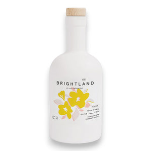 A white bottle of BRIGHTLAND's 'ARISE' Basil Infused Olive Oil comes with a natural wooden cap. The label features a bright yellow and white floral design, boasting 12.7 fl oz (375 mL) of 100% basil-infused olive oil. Enjoy its rare, bold, verdant taste that echoes California's vibrant essence, reminiscent of Peter Som's chic aesthetic.