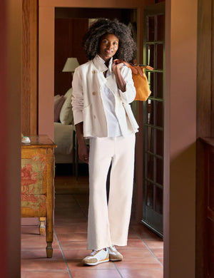 In a warmly lit hallway, a woman with curly hair wears the ultra-sophisticated FRANK & EILEEN - BELLA Italian Sweatpant in Vintage White, featuring an adjustable drawstring waist. She holds a brown bag over her shoulder and stands near elegantly designed wooden furniture in front of a glass-paned door.