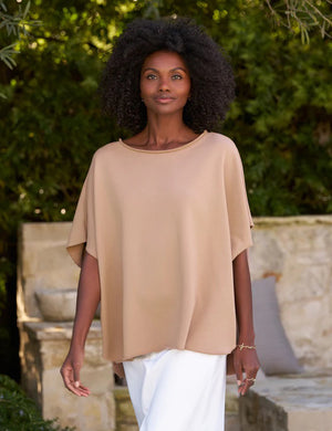 A woman with voluminous curly hair walks outdoors. She is wearing a loose-fitting FRANK AND EILEEN - OLIVE ORIGINAL CAPELET and white trousers, reminiscent of an original capelet. Behind her are lush green trees and a stone wall with a cushioned bench. She has a calm and confident expression.