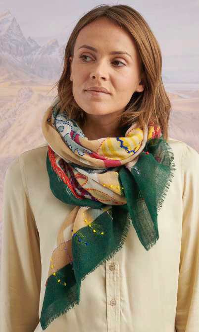 A person holds up the STORIATIPIC - ANDRES WOOL SCARF, an elegant rectangular accessory by STORIATIPIC, featuring colorful potted plants and vases with an orange border against a blurred natural backdrop.