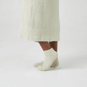 A person stands on a white background, wearing a cozy, textured white robe, KASHWERE - ADULT SOCKS by KASHWERE, and white slippers. Only the lower half of the body is visible. The attire appears ultra-plush and comfortable, ideal for lounging or relaxation.