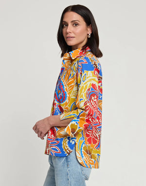 A woman with shoulder-length dark hair is shown from behind, wearing the HINSON WU Aileen 3/4 Sleeve Paisley Print Top, which features a colorful blend of red, yellow, blue, and orange shades. She has paired it with light blue denim jeans against a plain white background. This best-selling top from HINSON WU exudes style and vibrancy.