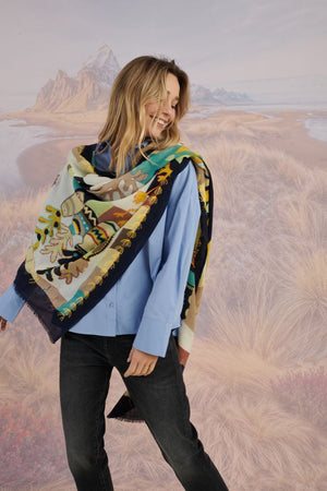 A woman with light brown hair wears a cream blouse and the STORIATIPIC - ANDRES WOOL SCARF by STORIATIPIC, elegantly hand-embroidered with green, yellow, red, and blue patterns. She gazes to the side against a scenic background.