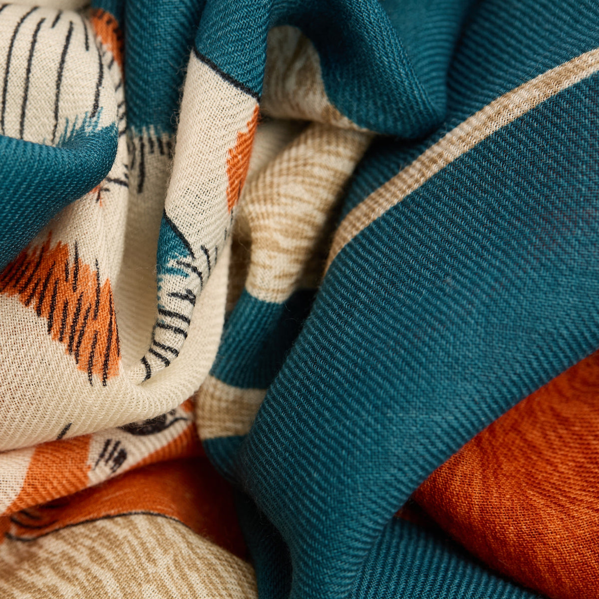 A woman with short blonde hair wears a dark jacket and the INOUI EDITIONS TOUTOU Dogs wool scarf in duck blue from INOUI, featuring playful animal illustrations in orange, teal, and cream, standing against a plain white background.
