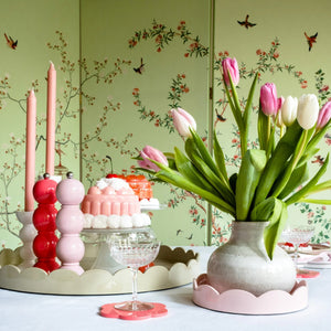 A table is set with colorful tulips in a vase, candles, and ornate dessert dishes featuring cakes. The background is a green wall with a delicate floral and bird pattern, creating a lively and charming ambiance. Adding to the charm are elegant ADDISON ROSS LONDON Bobbin Salt or Pepper Mills (24cm) by ADDISON ROSS alongside a watermelon pink mill.