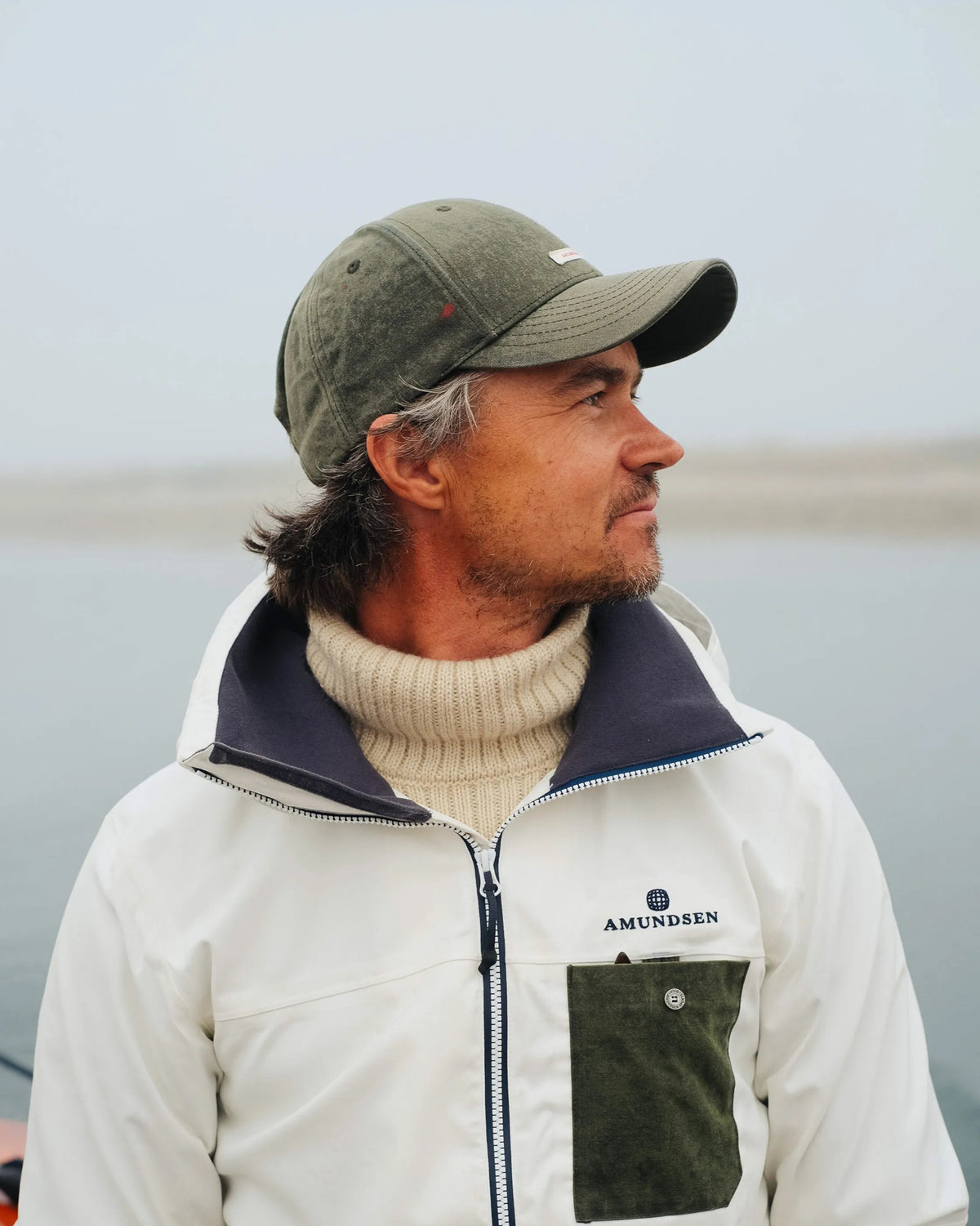 The AMUNDSEN SPORTS - WAXED COTTON CAP in navy blue is shown from the side against a plain white background, featuring a curved brim and an adjustable strap with a red, white, and black checkered norgesreima design.