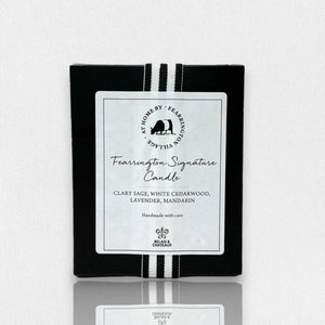 A rectangular black box prominently displays the label "Fearrington Signature Candle" at its center, reading "Clary Sage, White Cedarwood, Lavender, Mandarin." The top features a white circular logo with the subtitle "Handmade with care." As part of our luxurious natural soy candles collection, it is crafted by Fearrington Village - Home Decor and includes two vertical white stripes.