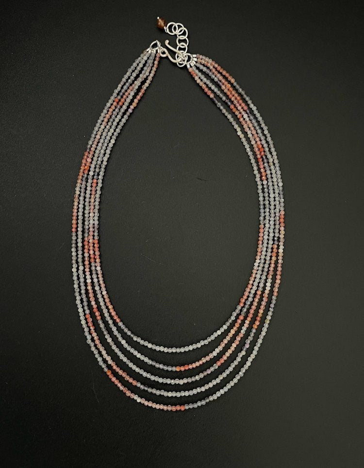 Introducing the ANN LIGHTFOOT - FIVE STRAND IOLITE AND CARNELIAN STRAND NECKLACE, a beautifully crafted piece showcasing multi-strand elegance with small beads in shades of red, grey, and white. The strands are gracefully layered, enhanced with carnelian accents, and secured with a silver clasp for a sophisticated finish.