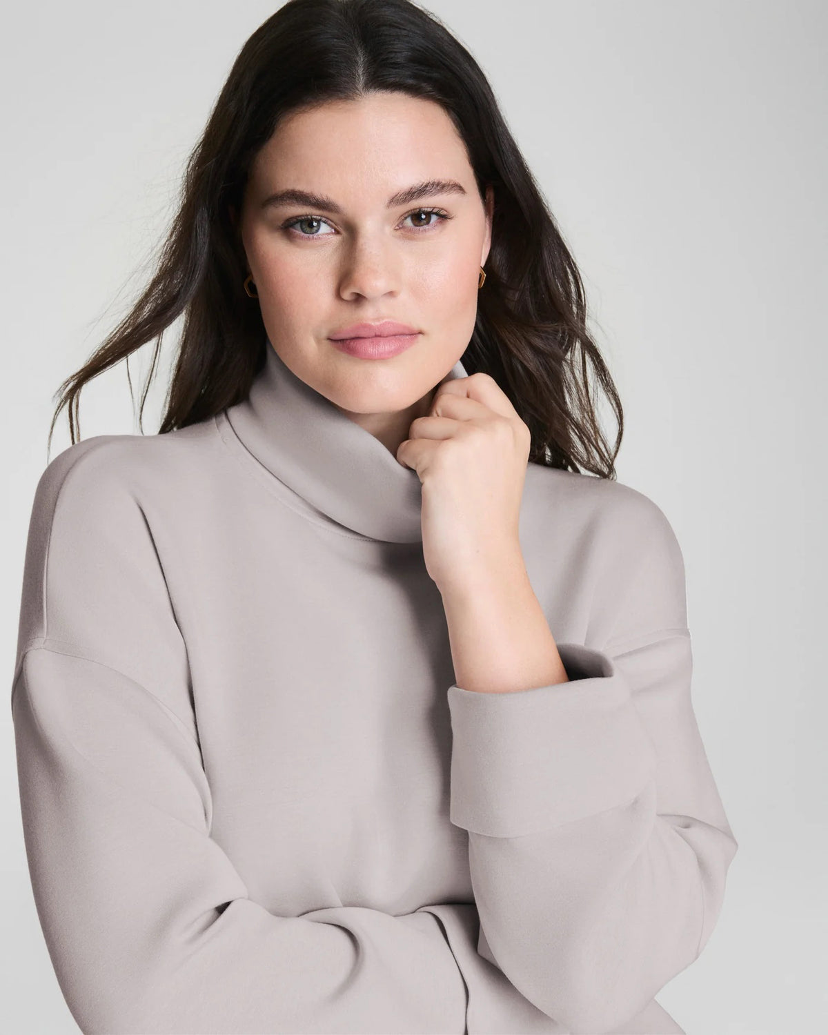 A woman confidently poses in the SPANX - AIRESSENTIALS TURTLENECK TOP and matching pants, crafted from ultra-drapey spacer fabric, against a plain light gray background with a slight smile.