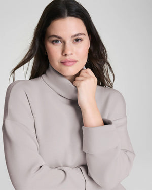 A woman confidently poses in the SPANX - AIRESSENTIALS TURTLENECK TOP and matching pants, crafted from ultra-drapey spacer fabric, against a plain light gray background with a slight smile.