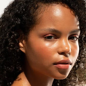 A close-up of a person with curly dark hair, a serious expression, and warm makeup featuring the universally flattering, weightless JASON WU - HOT FLUFF LIP/EYE/CHEEK by JASON WU BEAUTY on their face. They are looking directly at the camera with a neutral background.