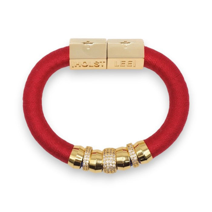 The HOLST + LEE - LUXE CLASSIC BRACELET by HOLST & LEE features an elegant orange cord with gold-plated accents, including a rectangular gold clasp adorned with several gold and diamond-encrusted beads near the clasp.