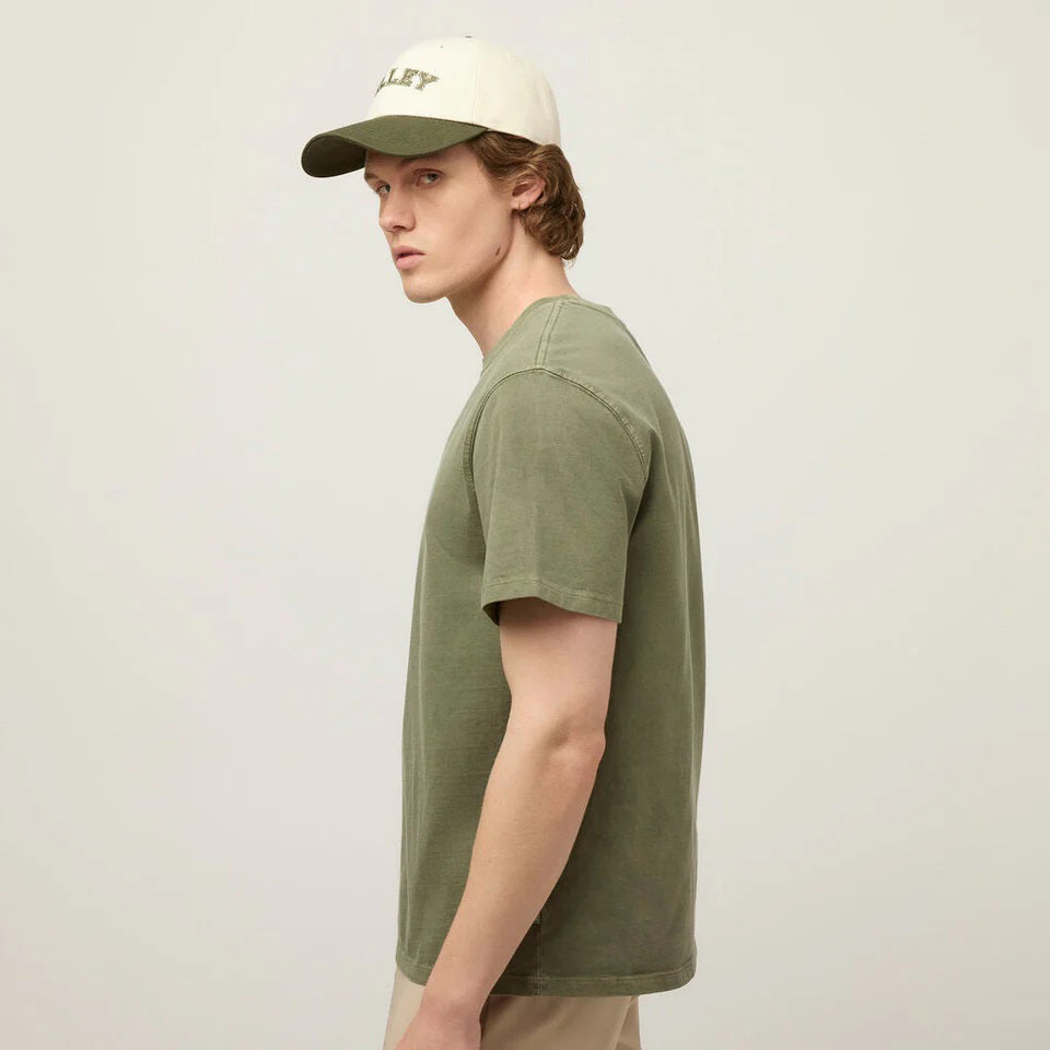 A young man with light skin and short brown hair wears the TILLEY Mid Weight Sunwashed T-Shirt in Thyme Green and a cream &quot;TILLEY&quot; baseball cap against a light, neutral background.