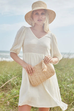 A person is standing on a grassy dune by the beach, adorned in a white dress and a straw hat, holding the MAR Y SOL - IVY CLUTCH featuring a crocheted raffia design. The ocean and sky form the backdrop of this serene scene.