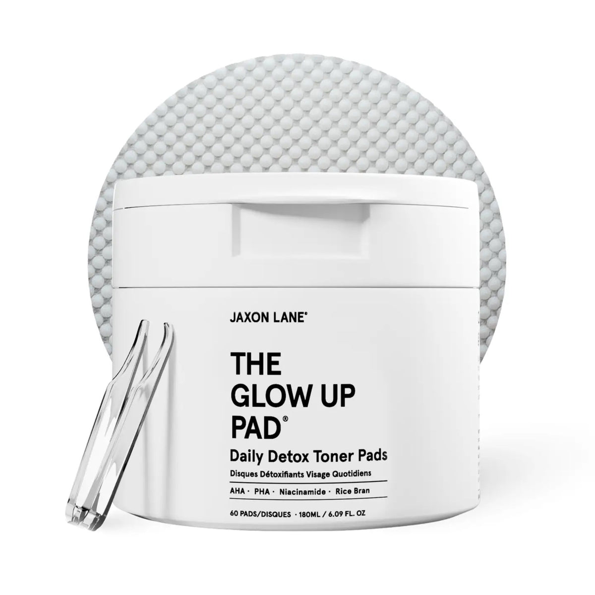 A white jar labeled JAXON LANE - GLOW UP PAD DAILY DETOX TONER PADS sits with a clear tweezer in front of a textured pad. These hydrating, biodegradable pads contain AHA, PHA, niacinamide, and rice bran.