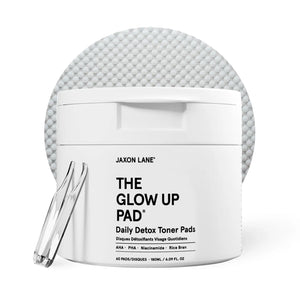 A white jar labeled JAXON LANE - GLOW UP PAD DAILY DETOX TONER PADS sits with a clear tweezer in front of a textured pad. These hydrating, biodegradable pads contain AHA, PHA, niacinamide, and rice bran.
